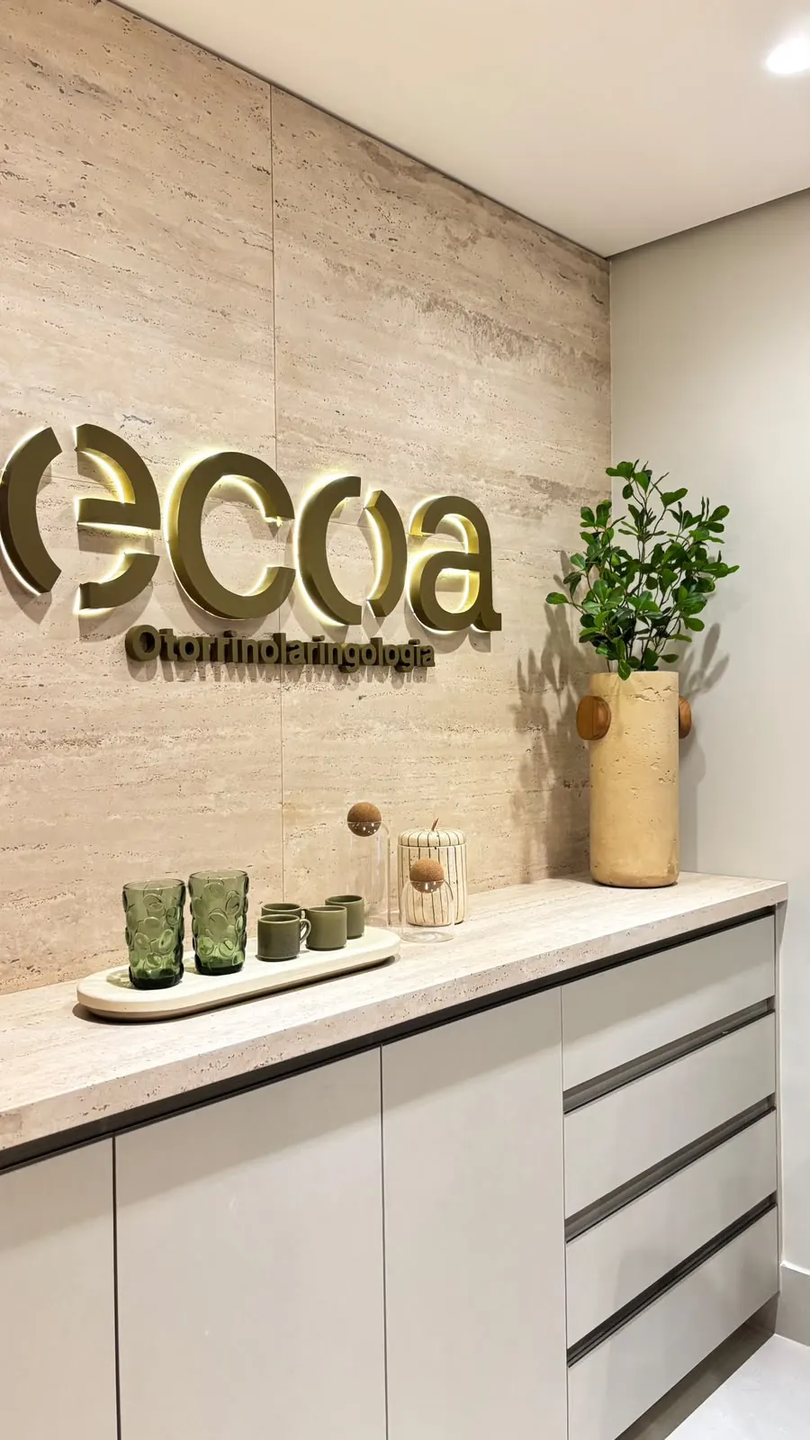 Recepção da ECOA Otorrinolaringologia, com o logotipo iluminado sobre a parede de travertino